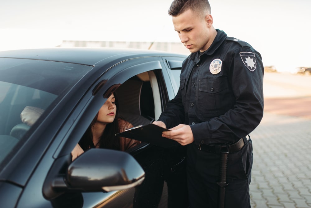 Police officer is reviewing a document while standing next to a female driver in a car during a traffic stop.
