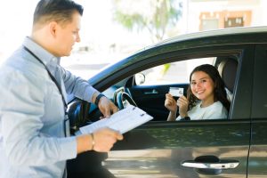 Smiling young driver holding a license while sitting in a car during a driving test with an instructor.