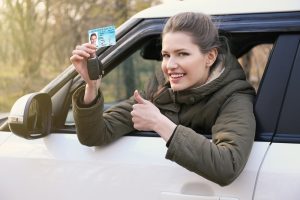 Woman smiling and holding a car key and driver’s license out of a vehicle window.