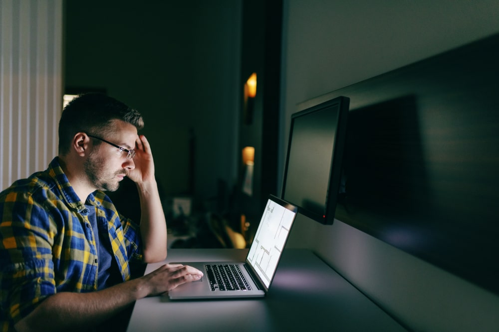 Worried man wearing glasses works on a laptop late at night, appearing stressed while reviewing something on the screen.