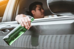 Driver holding a green beer bottle out the car window, suggesting drinking and driving.