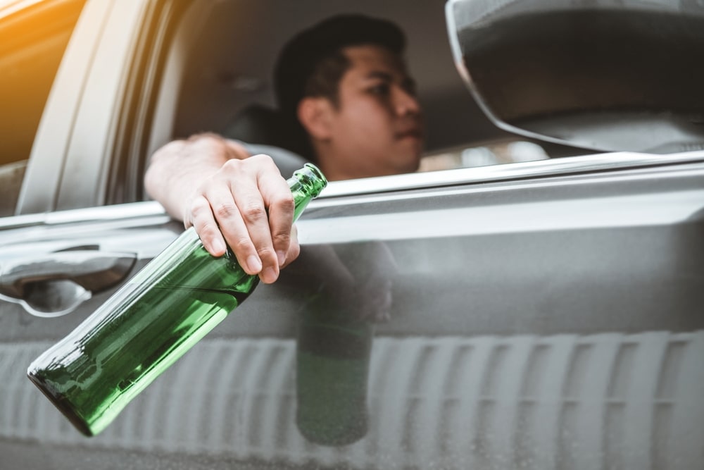 Driver holding a green beer bottle out the car window, suggesting drinking and driving.