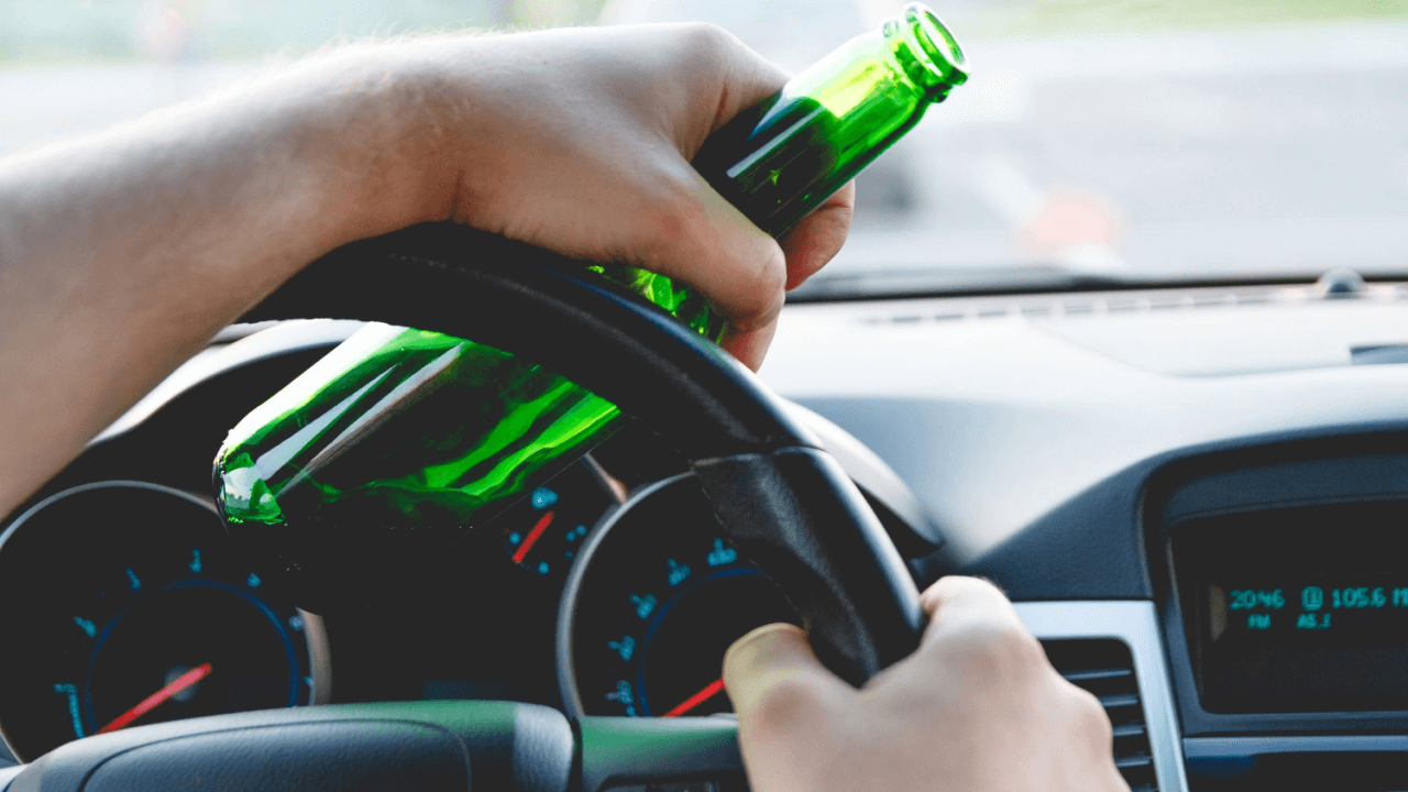 Person holding beer bottle behind steering wheel illustrating which state has the strictest laws for drunk driving